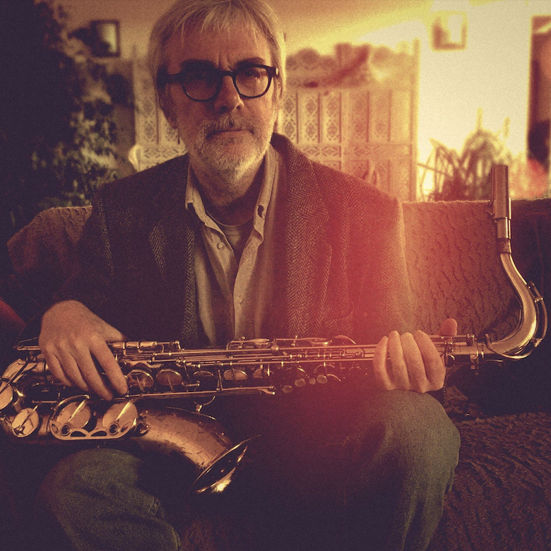 Jacques Dubuc, graphiste/directeur artistique, Verdié Voyages, 780x780 : Photo de profil sépia, avec reflet dégradé jaune à orange, de Jacques Dubuc en t-shirt, chemise, manteau en tweed et jean, assis sur un canapé et tenant un saxophone.