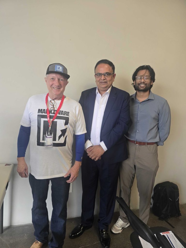 Trois participants debout ensemble à l'intérieur lors d'un événement professionnel, l'un d'eux portant un Markzware chemise et badge, posant pour une photo devant un mur neutre